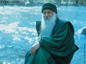 Osho in Green Robes and Knit Cap Seated by a Sparkling River HD wallpaper