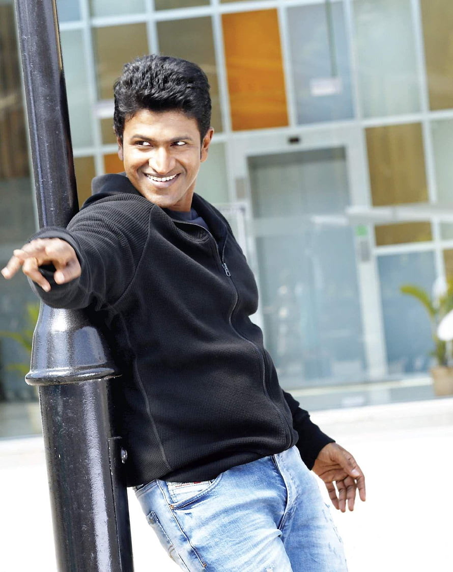 Puneeth Rajkumar smiling in black hoodie and jeans leaning on a pole HD phone wallpaper
