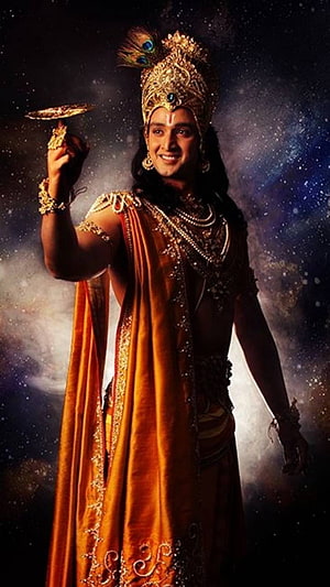 Krishna in ornate golden crown and saffron robes holding a discus against a cosmic starry background HD phone wallpaper