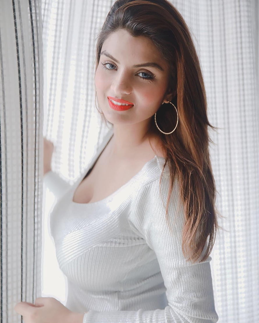 Anveshi Jain in a white ribbed top with hoop earrings standing by a window HD phone wallpaper