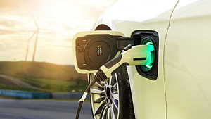 White Electric Vehicle Charging With Green Indicator Light And Wind Turbines In Background HD wallpaper
