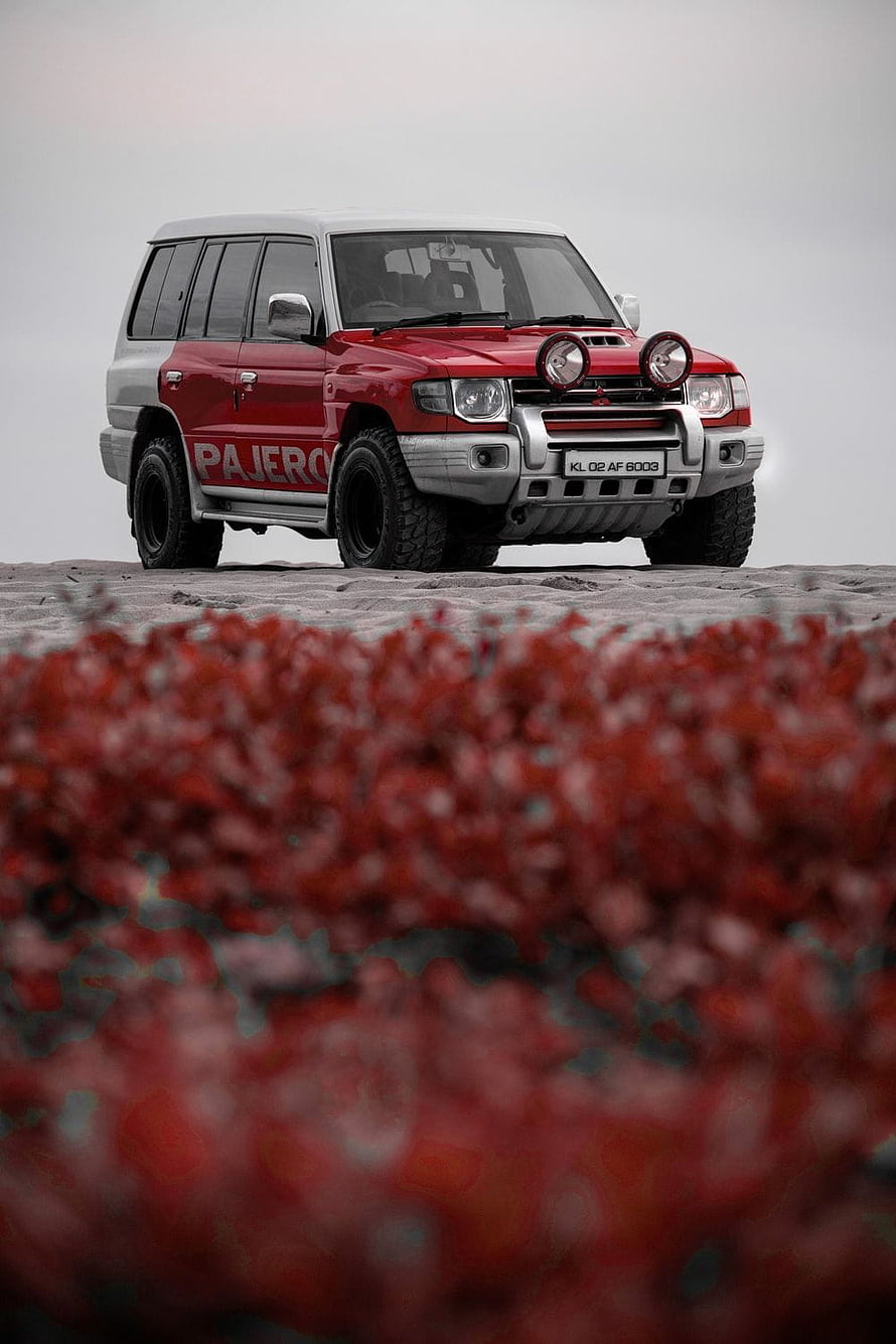 Red Mitsubishi Pajero SUV parked on a sandy beach with vibrant red foliage in the foreground HD phone wallpaper