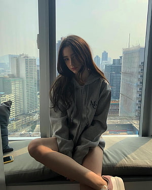 Dasha Taran in a grey hoodie sitting on a window seat with a city skyline view HD phone wallpaper