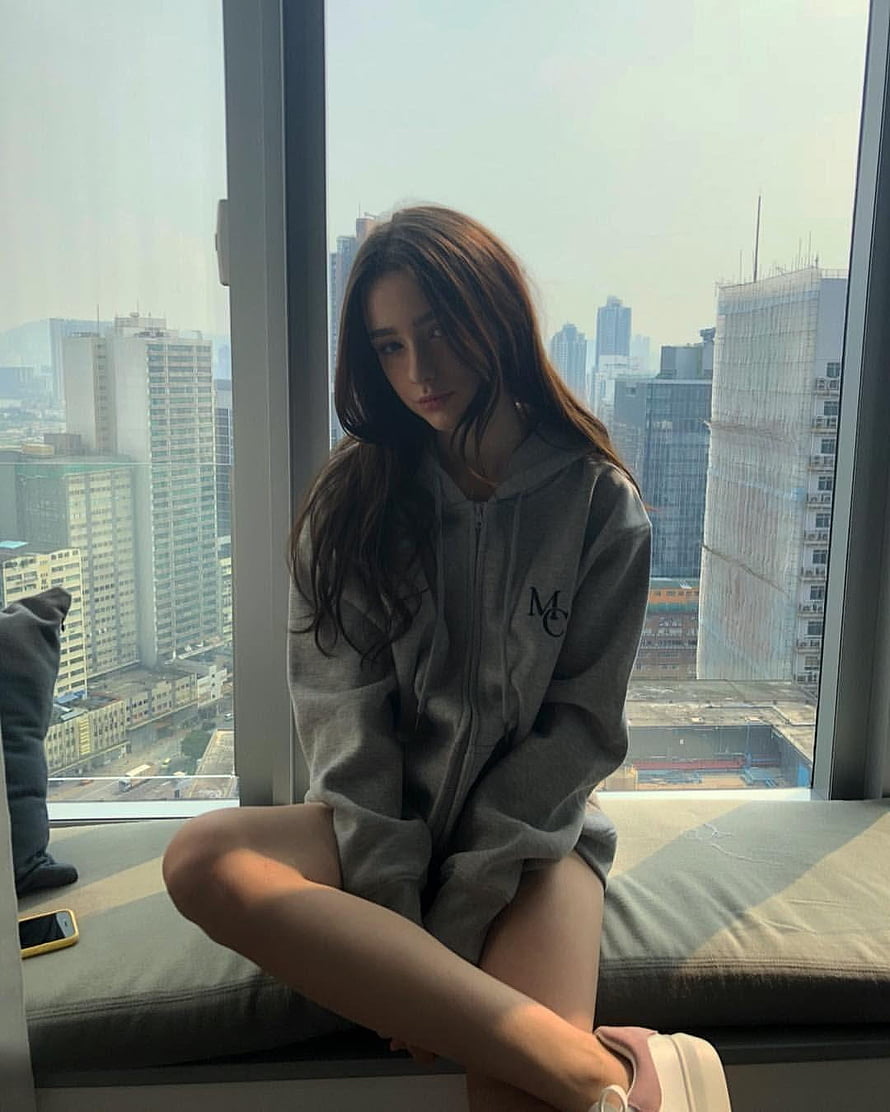 Dasha Taran in a grey hoodie sitting on a window seat with a city skyline view HD phone wallpaper