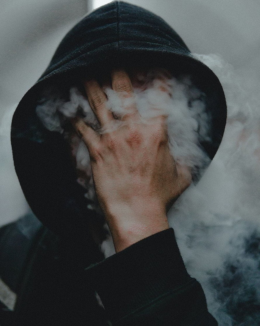 Mysterious hooded figure with hand covering face surrounded by swirling smoke HD phone wallpaper