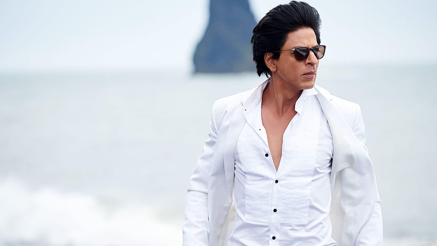 Shah Rukh Khan in a white suit and sunglasses standing on a misty beach with a rocky outcrop in the background HD wallpaper