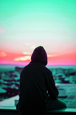 Silhouette of a hooded figure gazing at a vibrant sunset over a city skyline HD phone wallpaper