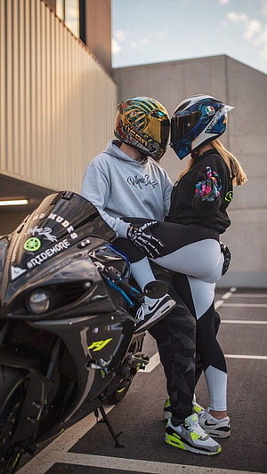 Couple in full-face helmets embracing beside a black sport motorcycle in a parking lot HD phone wallpaper