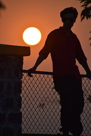 Silhouette of a man in sunglasses leaning on a railing against a vibrant orange sunset sky HD phone wallpaper