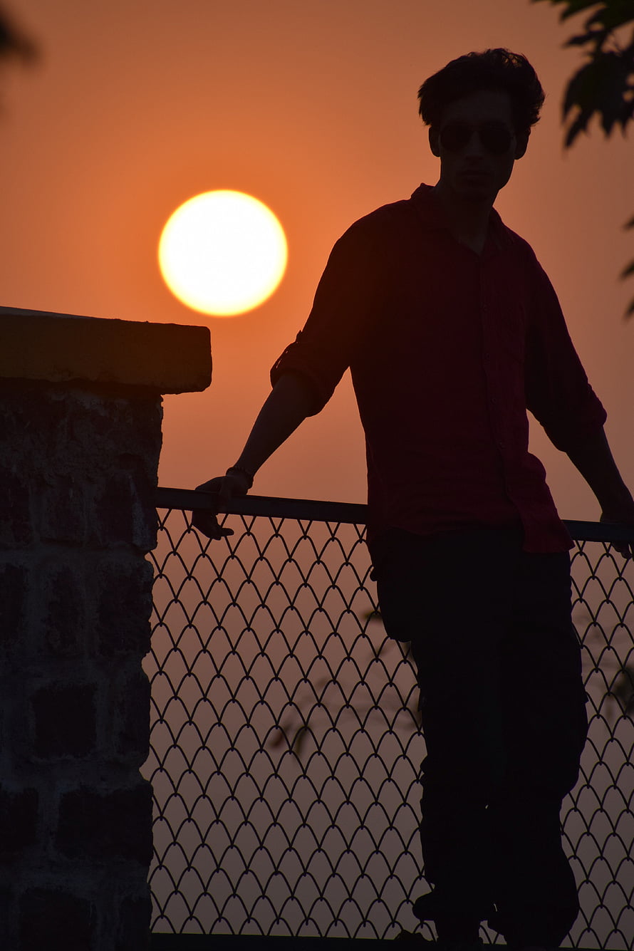Silhouette of a man in sunglasses leaning on a railing against a vibrant orange sunset sky HD phone wallpaper