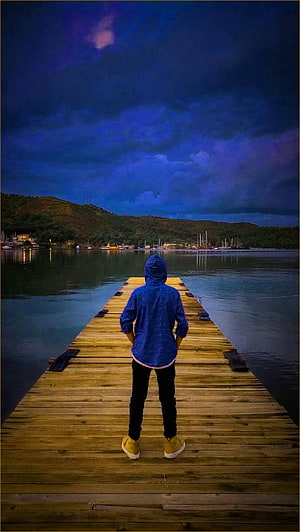 Lone figure in blue hoodie standing on wooden pier at dusk with moody storm clouds HD phone wallpaper