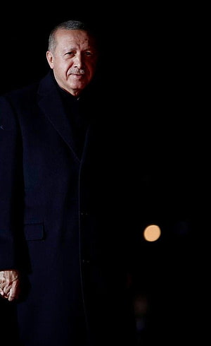 Recep Tayyip Erdoğan in a dark overcoat against a black background with a single warm bokeh light HD phone wallpaper