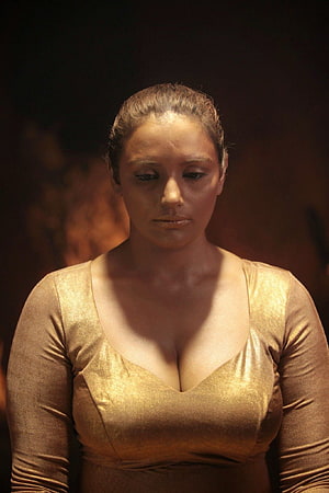Swetha Menon in a shimmering gold dress with a contemplative expression against a dark background HD phone wallpaper