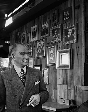 Mustafa Kemal Atatürk in a vintage suit holding a cigarette in front of a wooden wall with framed portraits HD phone wallpaper