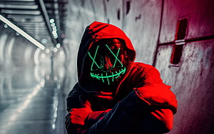 Figure in red hoodie wearing a glowing green neon mask in a dim tunnel HD wallpaper