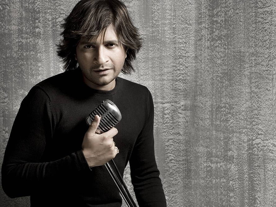 A man in a black sweater holding a vintage microphone against a textured gray wall HD wallpaper