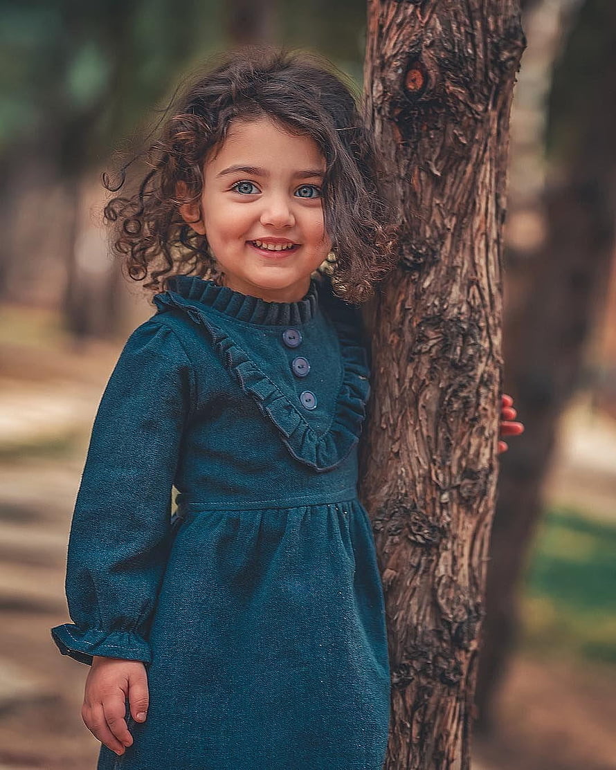 Smiling Curly-Haired Girl in Teal Dress Leaning Against Tree Trunk in Forest HD phone wallpaper