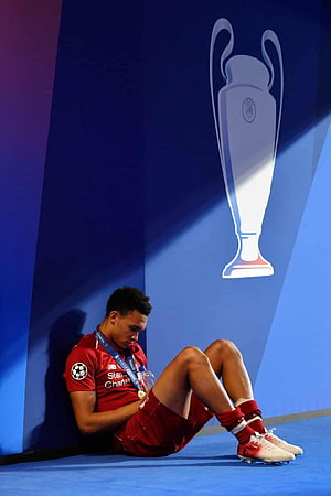 Trent Alexander-Arnold in red Liverpool kit sitting on pitch with Champions League trophy backdrop HD phone wallpaper
