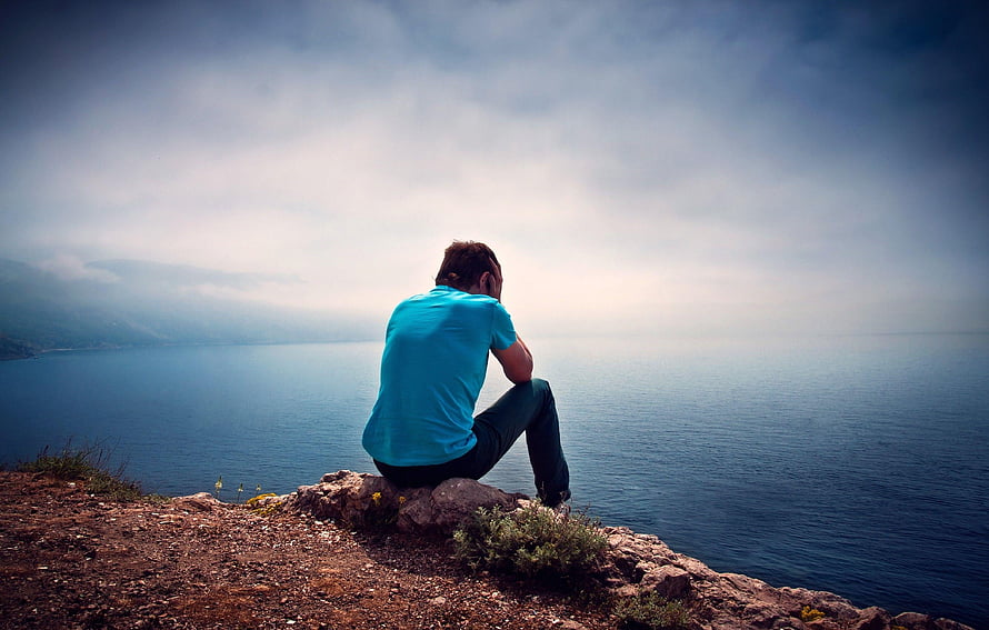 Man sitting alone on a rocky cliff overlooking a vast misty lake under a cloudy sky HD wallpaper