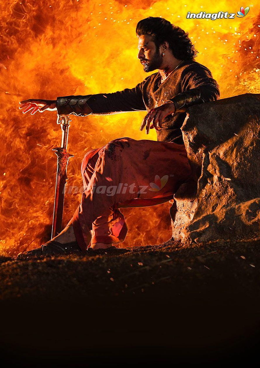 Baahubali warrior seated on rock with sword against fiery orange explosion backdrop HD phone wallpaper