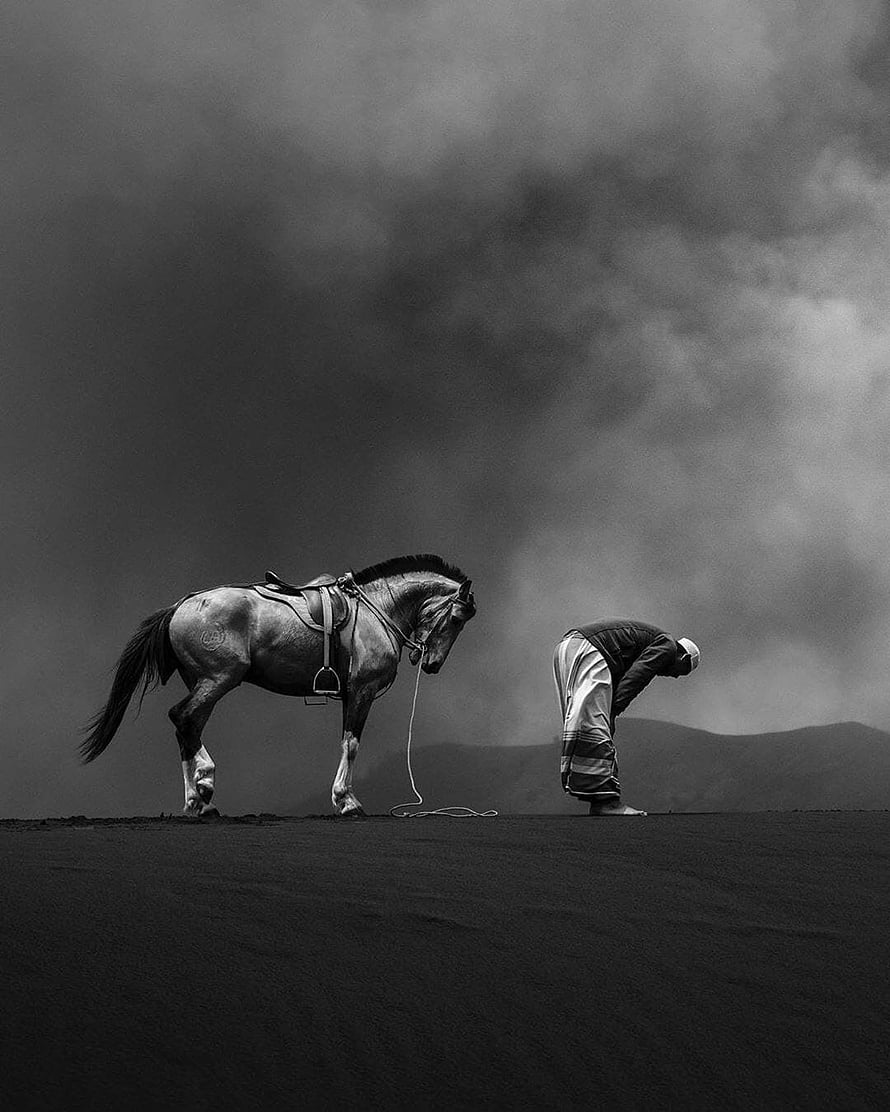 Black and White Photograph of a Man in Sujud Prayer Beside a Saddle Horse Under a Dramatic Cloudy Sky HD phone wallpaper