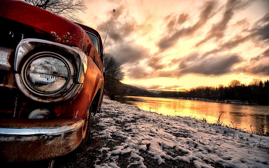 Rusty vintage car parked on a snowy riverbank under a dramatic golden sunset sky HD wallpaper