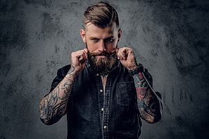 Man with full beard and sleeve tattoos posing against dark textured wall HD wallpaper