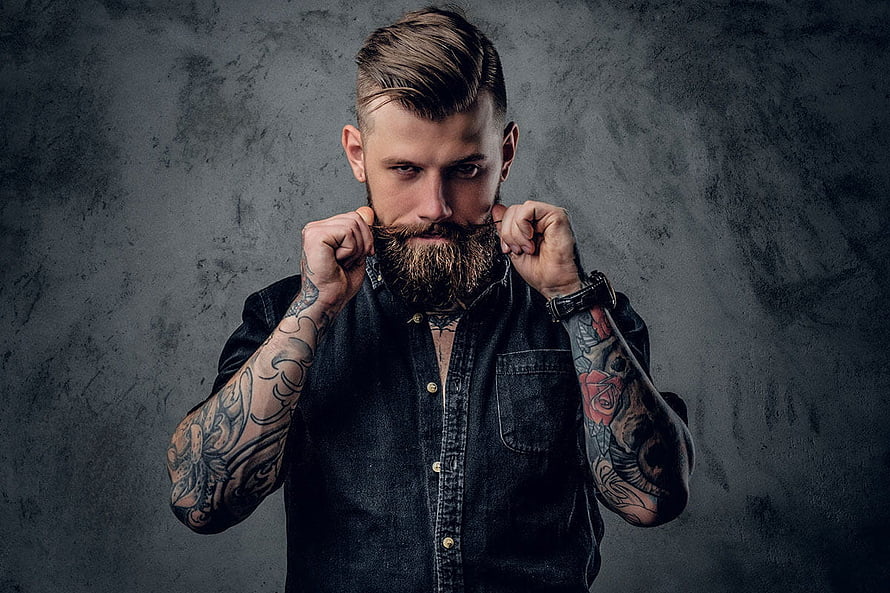 Man with full beard and sleeve tattoos posing against dark textured wall HD wallpaper