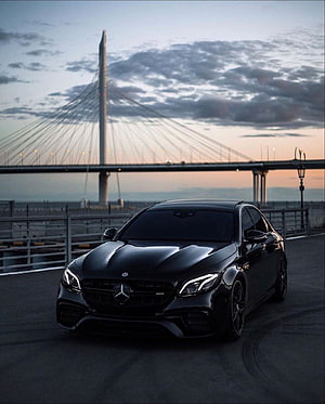 Black Mercedes-AMG E63 S Coupe Parked on a Coastal Road at Dusk with a Cable-Stayed Bridge in the Background HD phone wallpaper