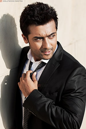 Suriya in a black suit adjusting his tie against a sunlit wall HD phone wallpaper