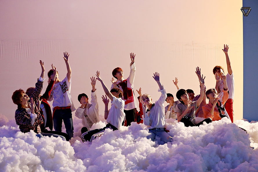 Seventeen members posing on a cloud set with raised hands in a pastel-toned studio HD wallpaper