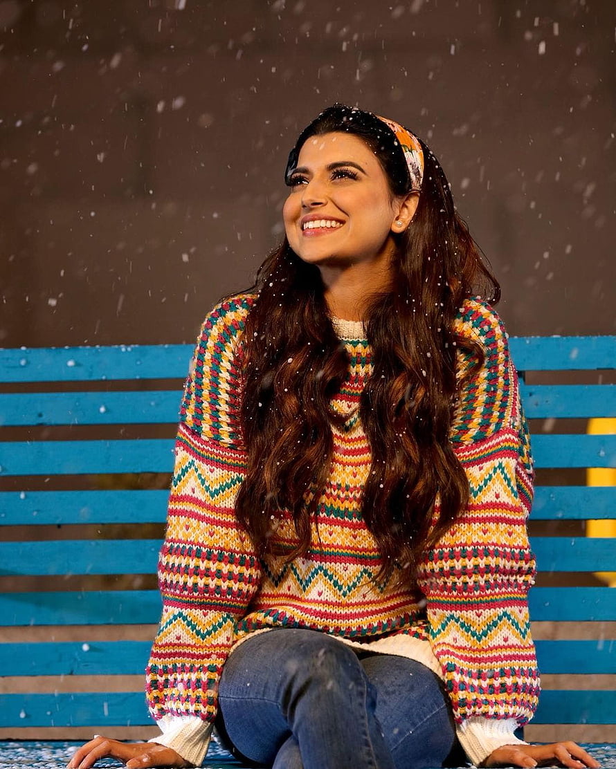 Nimrat Khaira in a colorful knit sweater sitting on a blue bench in falling snow HD phone wallpaper