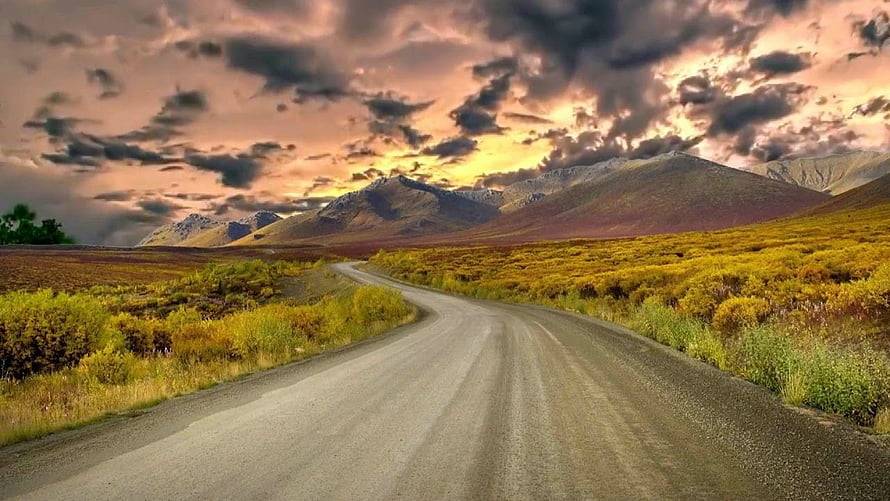 Winding gravel road through golden tundra hills under a dramatic sunset sky HD wallpaper