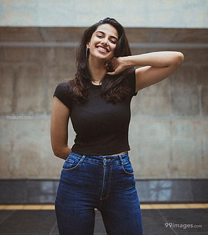 Meenakshi Chaudhary smiling in a black top and blue jeans against a concrete wall HD phone wallpaper