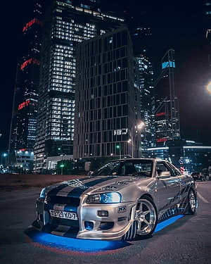 Silver Nissan Skyline R34 GT-R with Blue Underglow Parked in a City at Night HD phone wallpaper