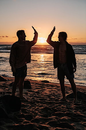 Two friends high-fiving on a sandy beach at sunset with ocean waves in the background HD phone wallpaper
