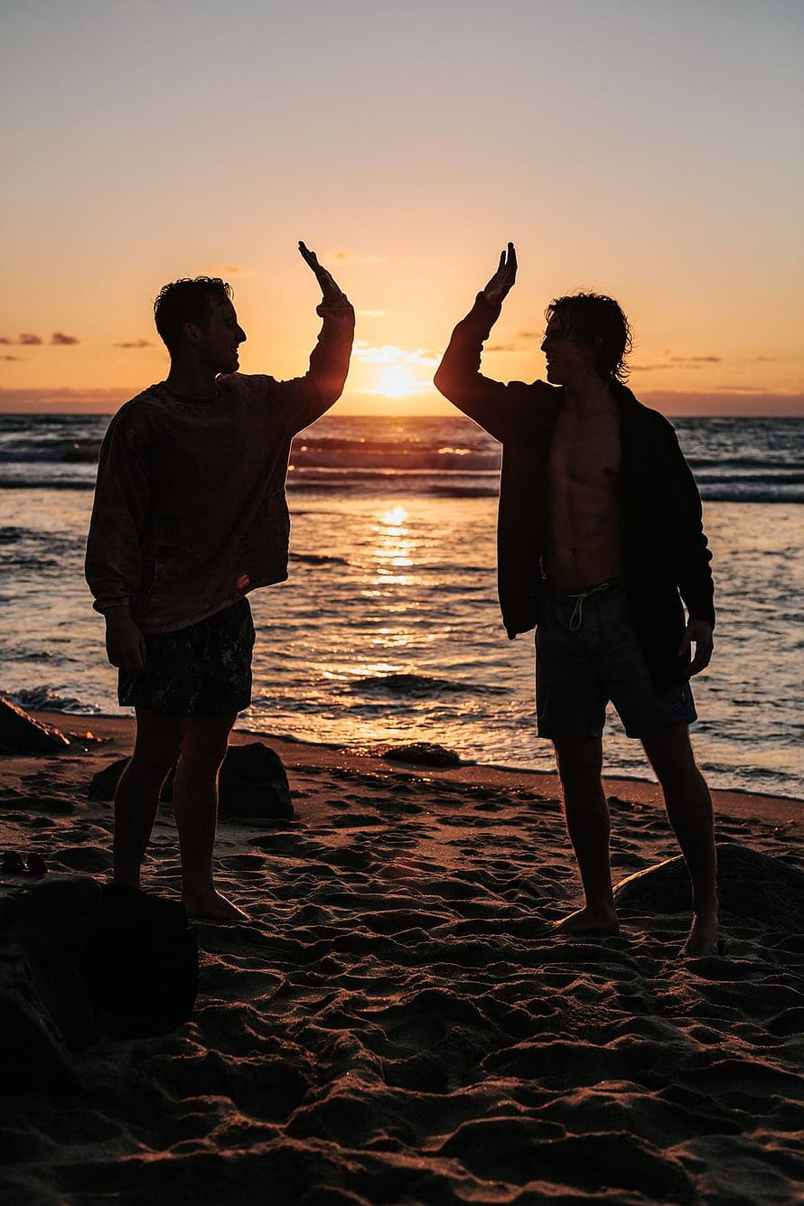 Two friends high-fiving on a sandy beach at sunset with ocean waves in the background HD phone wallpaper
