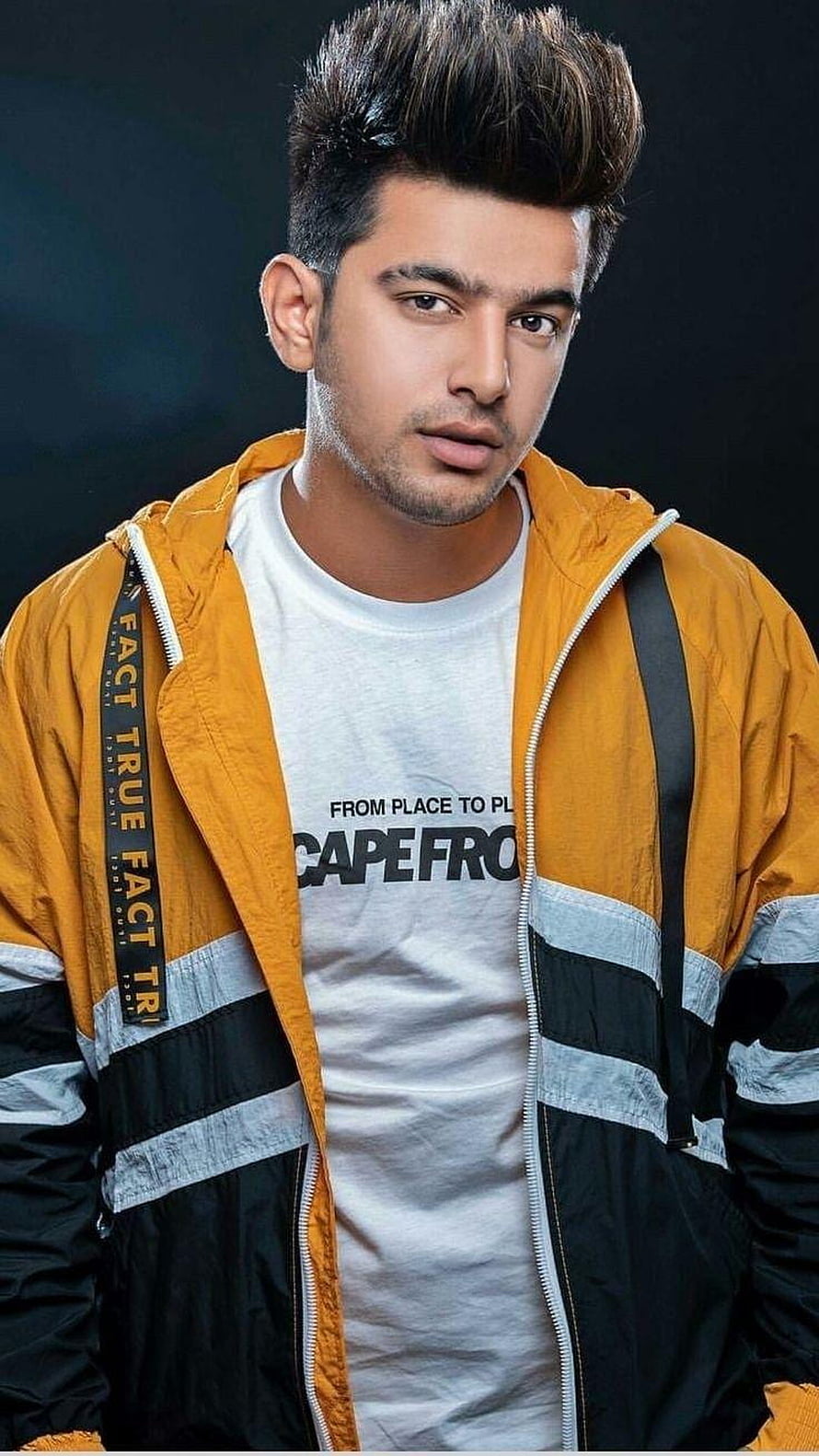 Jass Manak in a yellow and black jacket with a white graphic t-shirt against a dark studio background HD phone wallpaper