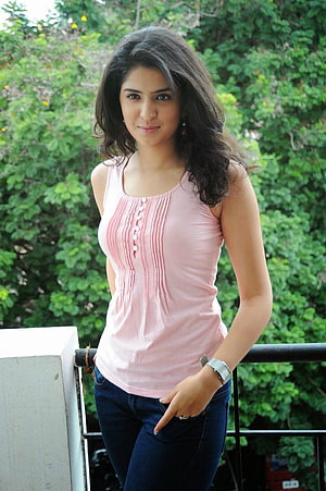 Young woman in a pink sleeveless top and dark jeans standing on a balcony with lush green foliage in the background HD phone wallpaper
