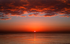 Vibrant red sunset over a calm ocean with dramatic clouds and a glowing horizon HD wallpaper