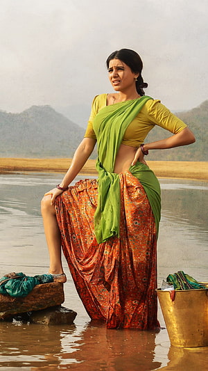 Samantha in a green and orange sari standing in a river with a brass bucket and laundry HD phone wallpaper