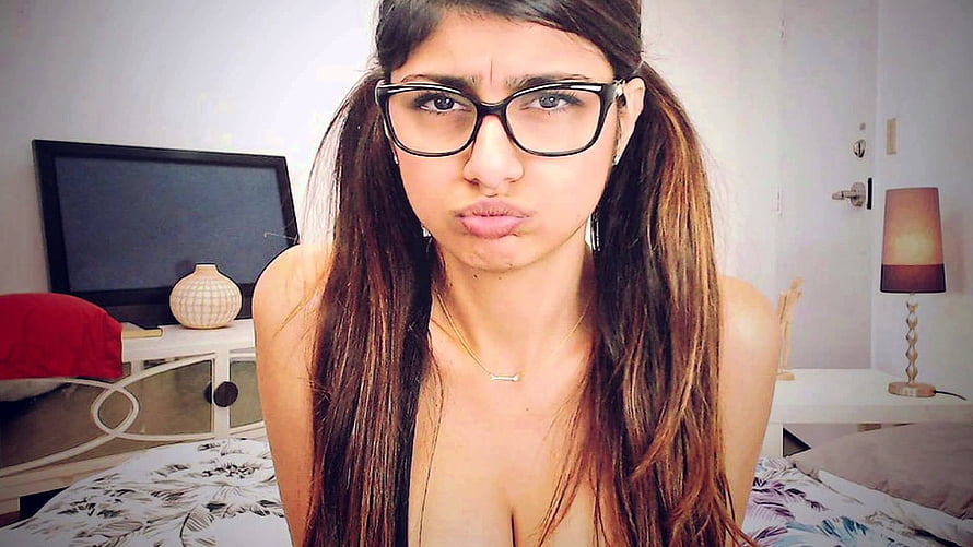 Young woman with long dark hair and black-rimmed glasses making a playful pouty expression in a bedroom HD wallpaper