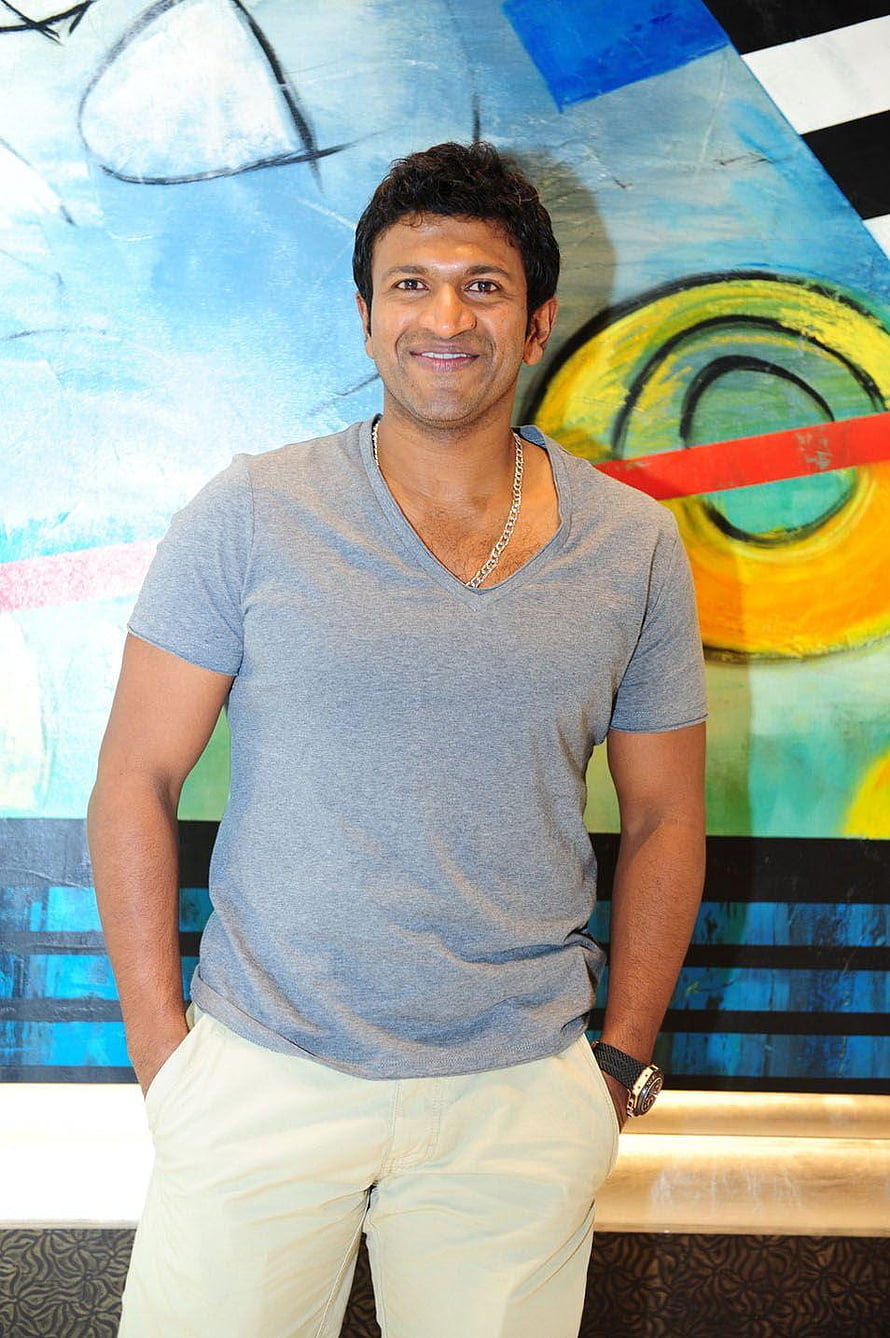Puneeth Rajkumar in a grey V-neck t-shirt and light trousers posing in front of a colorful abstract painting HD phone wallpaper