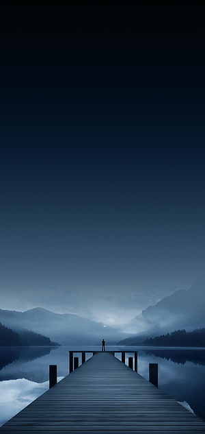 Solitary Figure Standing on a Wooden Pier Over a Misty Mountain Lake at Dusk HD phone wallpaper
