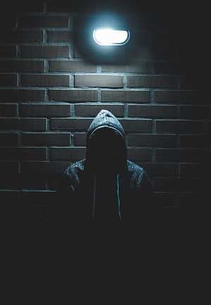 Mysterious hooded figure standing in shadow beneath a single light against a dark brick wall HD phone wallpaper