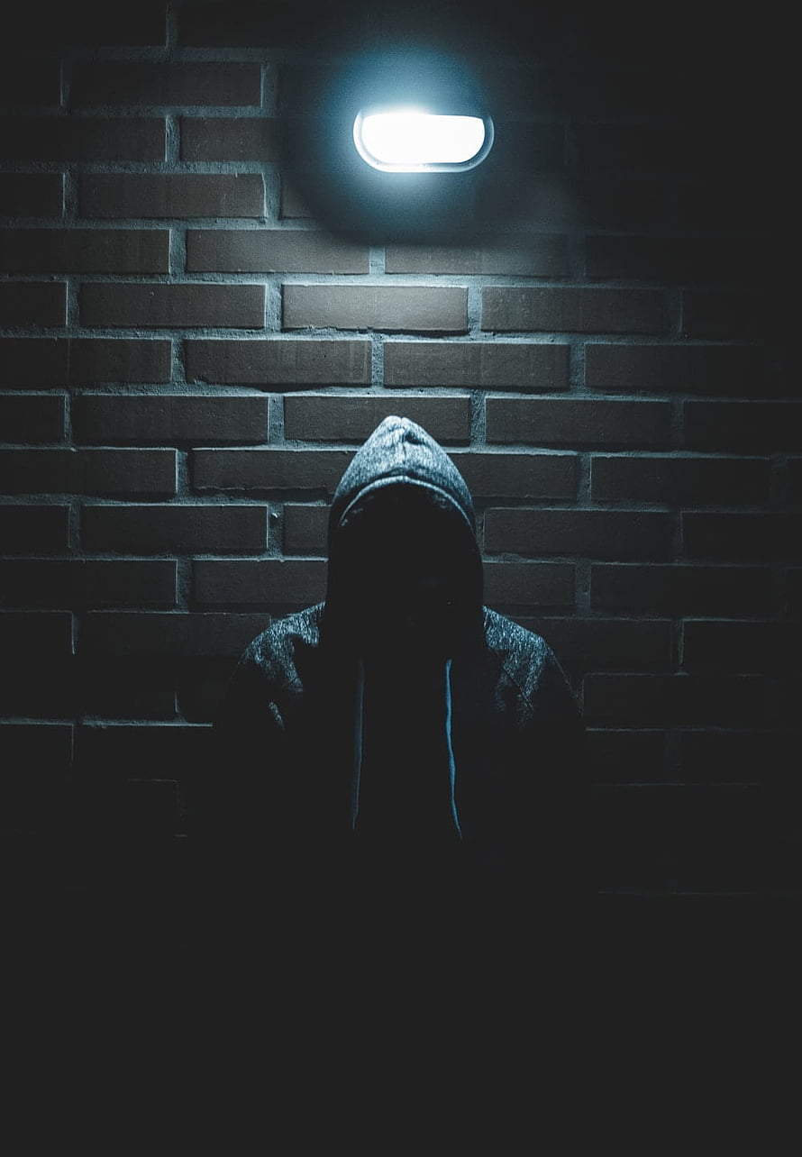 Mysterious hooded figure standing in shadow beneath a single light against a dark brick wall HD phone wallpaper