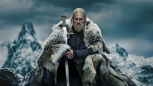 Bjorn Ironside from Vikings seated on a rocky outcrop in a fur cloak holding a sword against snowy mountains HD wallpaper