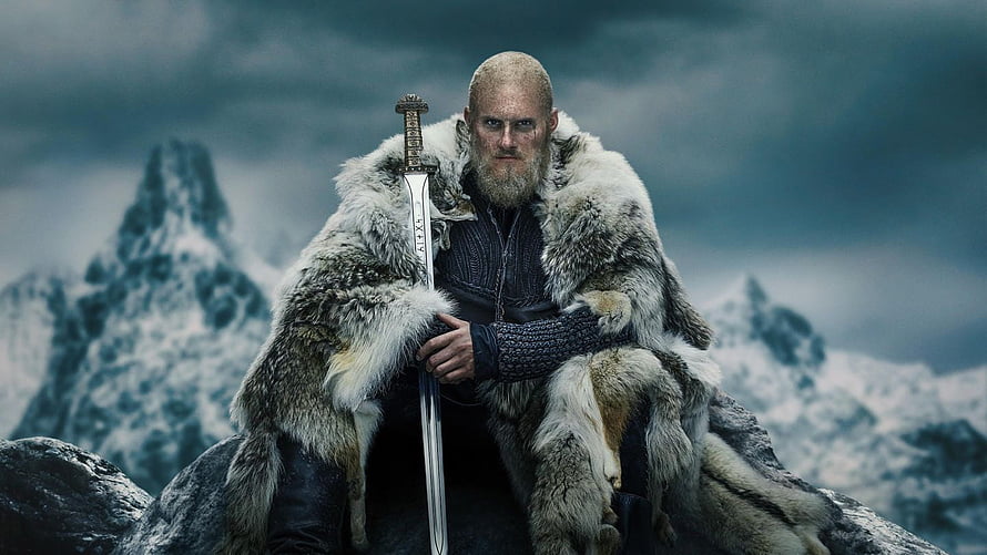 Bjorn Ironside from Vikings seated on a rocky outcrop in a fur cloak holding a sword against snowy mountains HD wallpaper