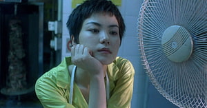 Chungking Express cinematic still of a pensive woman in a yellow shirt beside a vintage fan HD wallpaper