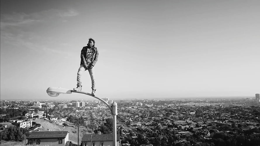 Kendrick Lamar standing on a street lamp overlooking a sprawling cityscape in black and white HD wallpaper